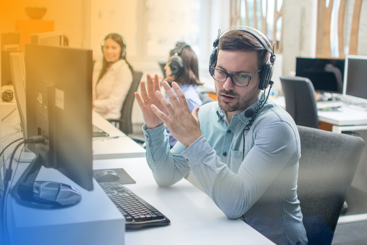 A customer service representative wearing a headset and glasses communicates with a client at a contact center, with colleagues working at computers in the background.
