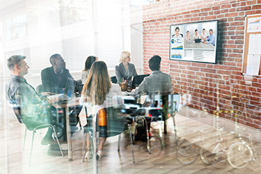 A group of business professionals are sitting around a conference table in a modern office, participating in a video conference displayed on a large monitor. The room features exposed brick walls and large windows, creating a bright and collaborative workspace.