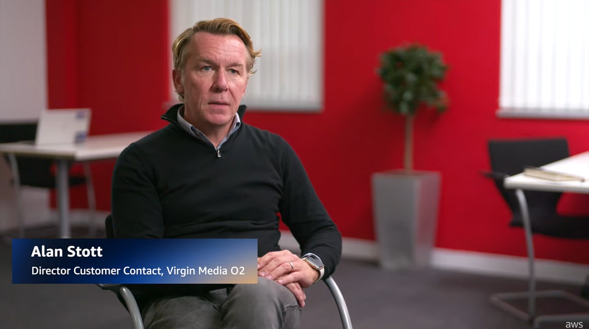 A person sits in a modern office setting, speaking about transforming customer conversations for Virgin Media O2. The background includes a red wall, white window blinds, a potted plant, and office furniture.