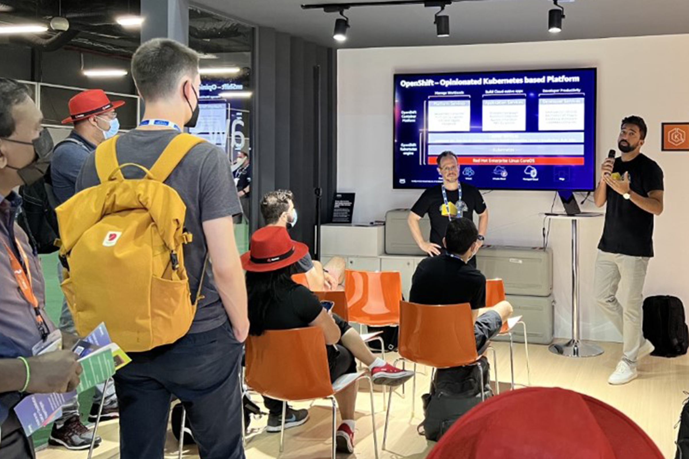A group of attendees watch an AWS speaker present on OpenShift, an opinionated Kubernetes-based platform, at a KubeCon event. The presentation is shown on a large screen, and participants are seated or standing, some wearing event badges and branded hats.