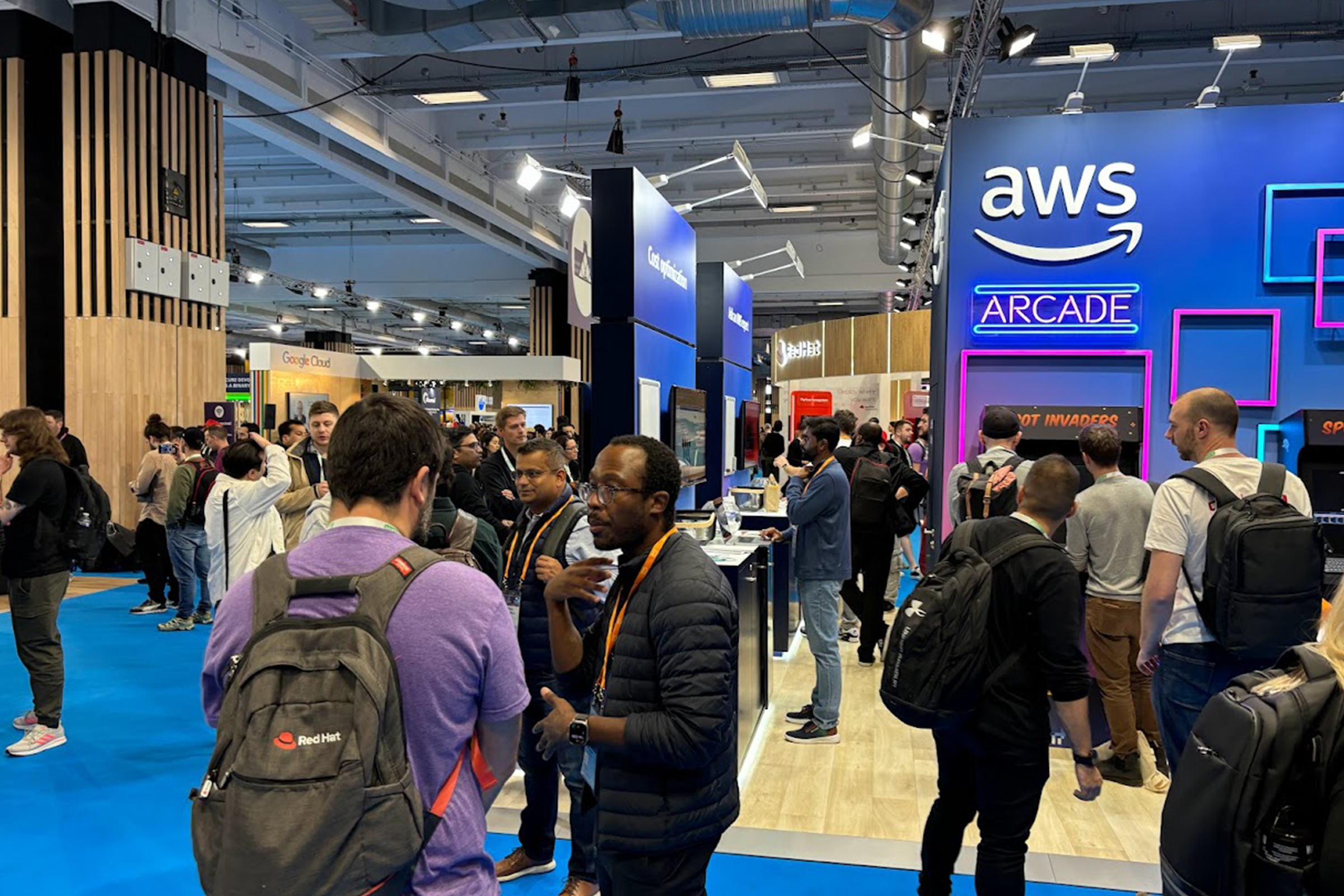 People interact and network at the AWS Arcade booth during the KubeCon event, with vibrant lights and displays in a busy exhibition hall.