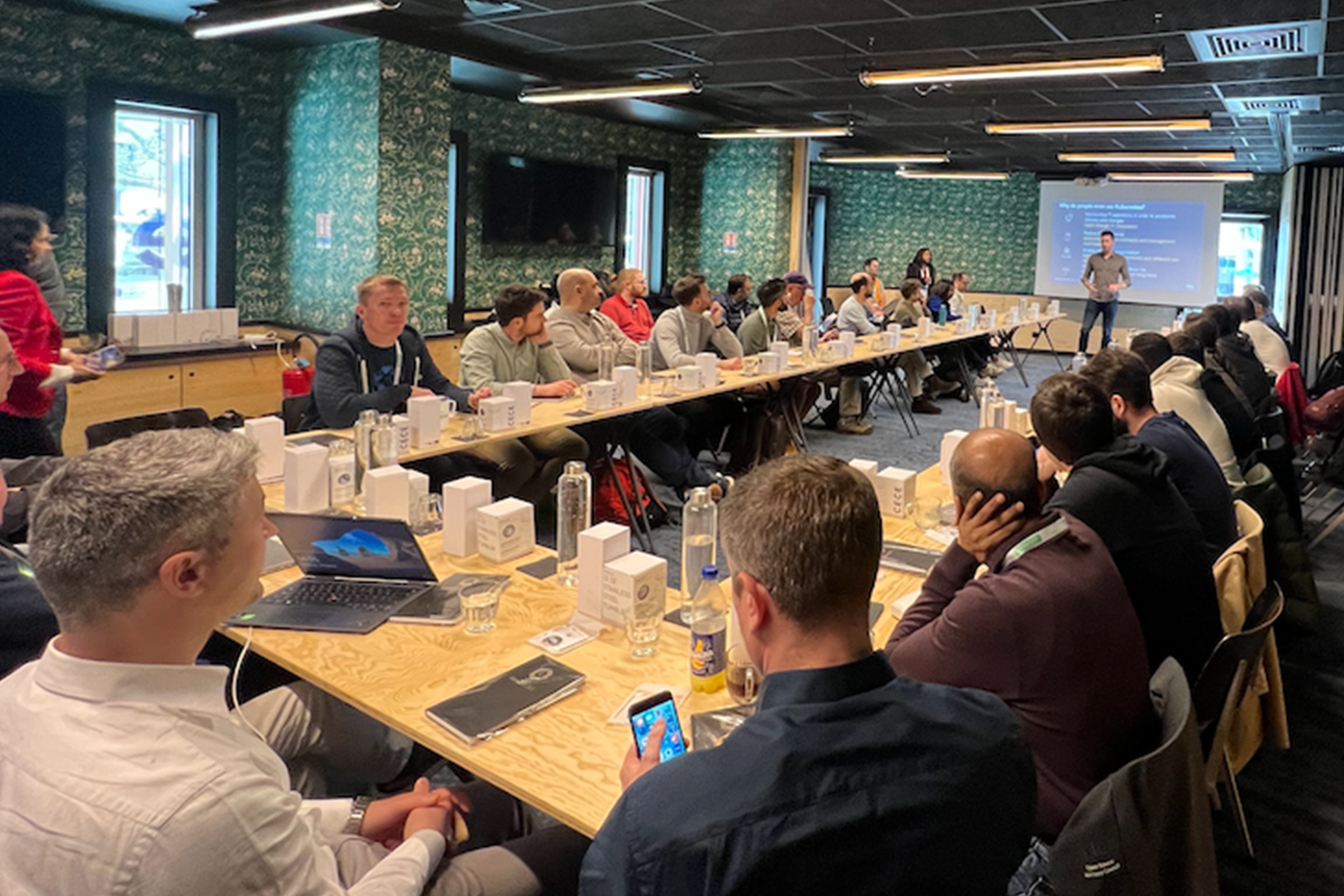 A group of people attending an AWS Retail workshop or conference session at KubeCon, sitting around a large U-shaped table, listening to a presenter at the front of the room.