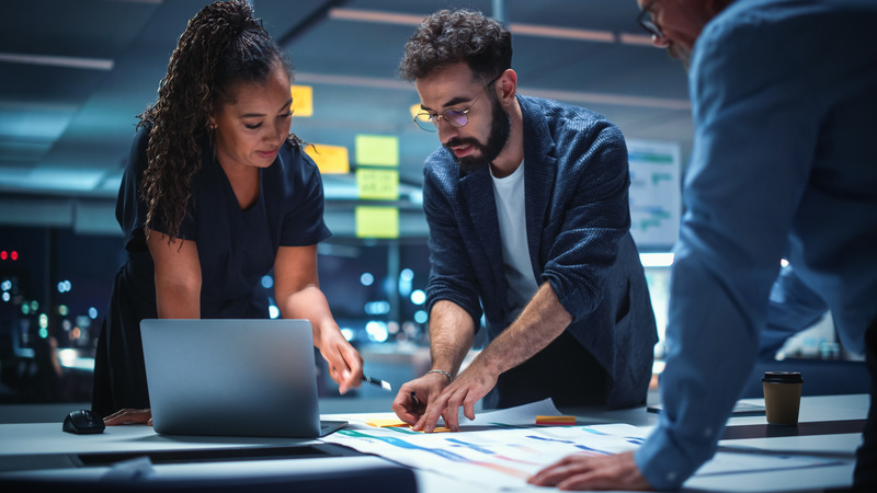 Three colleagues collaborate on a project in a modern office at night, working together around a laptop and documents on a desk.