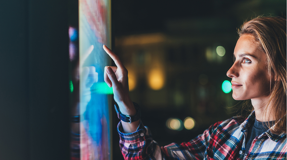 A woman interacts with a large illuminated digital touchscreen display in an urban nighttime setting.