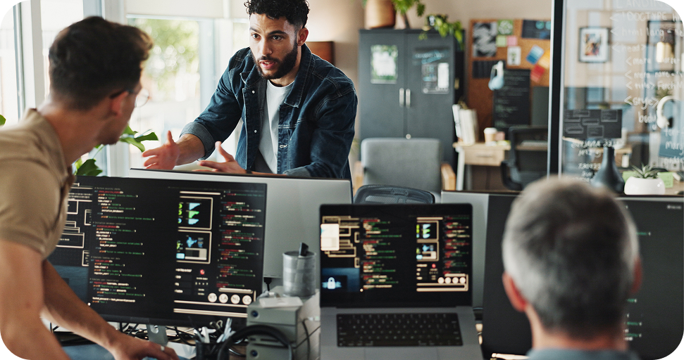 Three software developers collaborating around multiple monitors displaying code in a modern office workspace with natural lighting and plants.