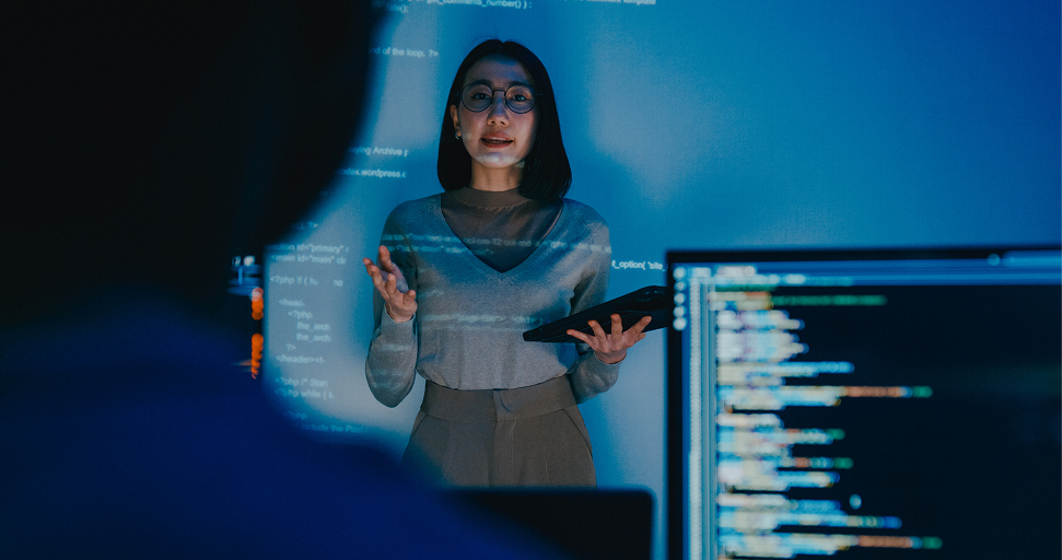 A female developer wearing glasses gestures while presenting code projected on screens in a blue-lit modern tech workspace.