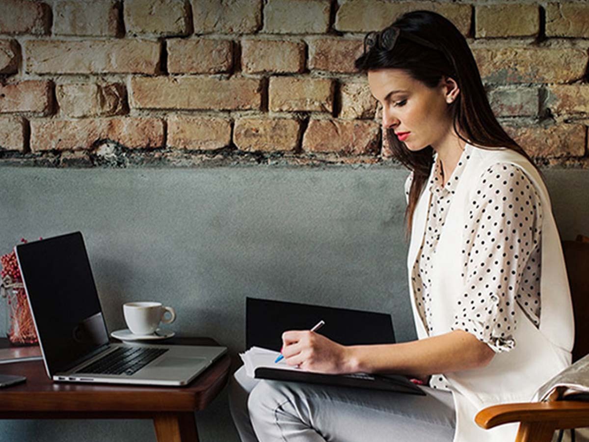 A woman sitting at a table in a cafe, working on a laptop and writing in a notebook. There is a cup of coffee on the table and a brick wall in the background.