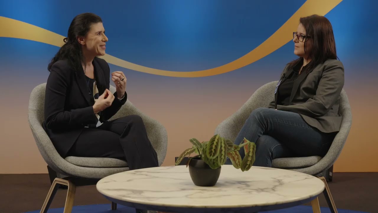 Two women having an interview, sitting in modern chairs with a round table and plant between them, in a contemporary setting with a blue and orange background.