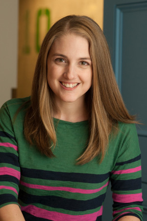 A woman with long straight hair, wearing a green sweater with colorful stripes, smiling indoors near a blue door and a wall with the number 10 in the background.