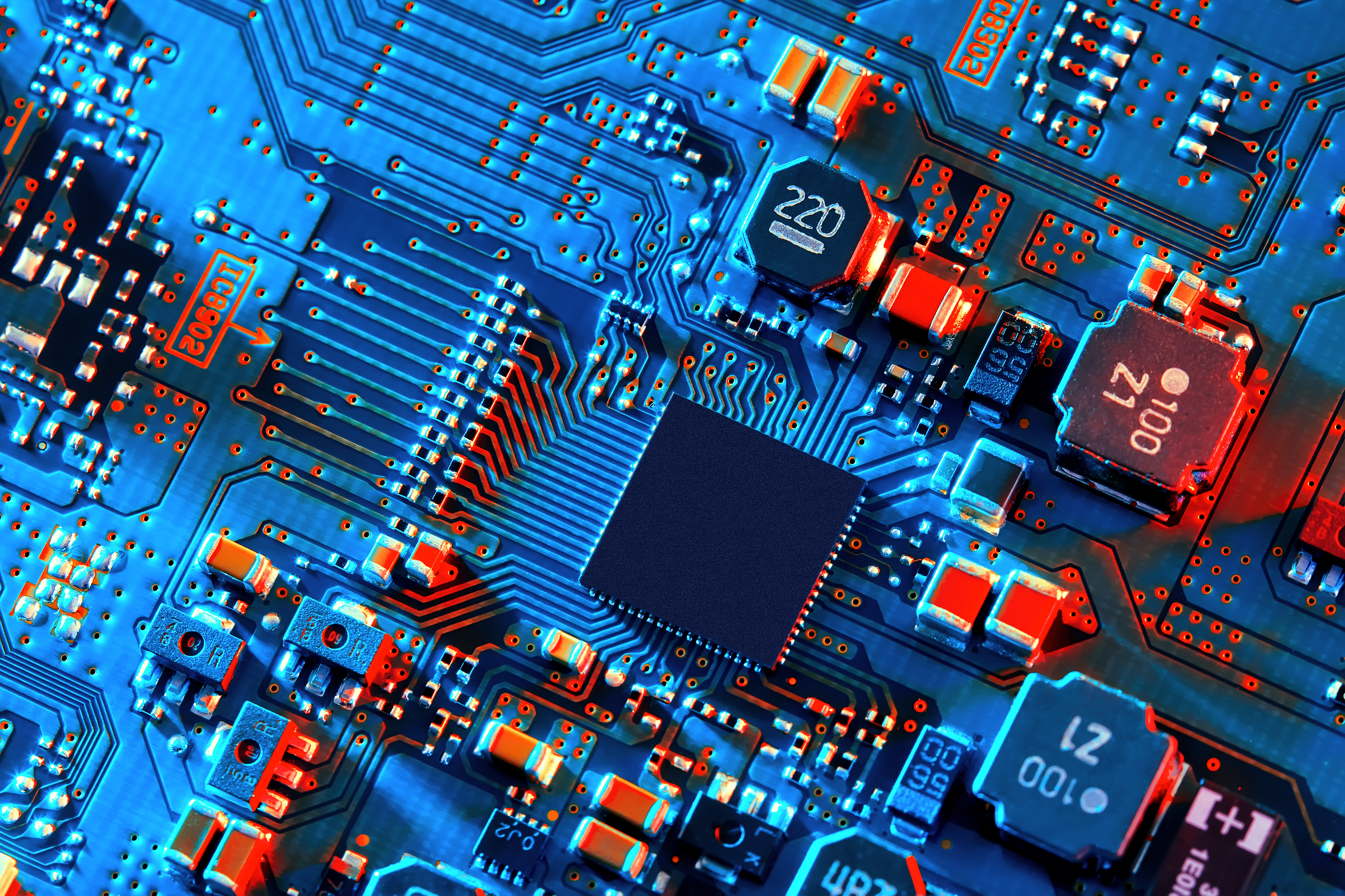 Close-up of a blue circuit board with a central microchip and various electronic components illuminated by red and blue lighting.