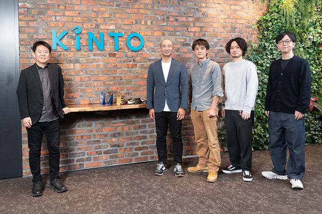 A group photo of the KINTO team standing in front of a brick wall with the KINTO logo, featuring five people in casual business attire.