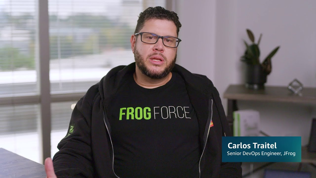 A person wearing a black "FROG FORCE" shirt and jacket, seated in an office with a plant and shelves in the background; text overlay reads "Carlos Traitel, Senior DevOps Engineer, JFrog."