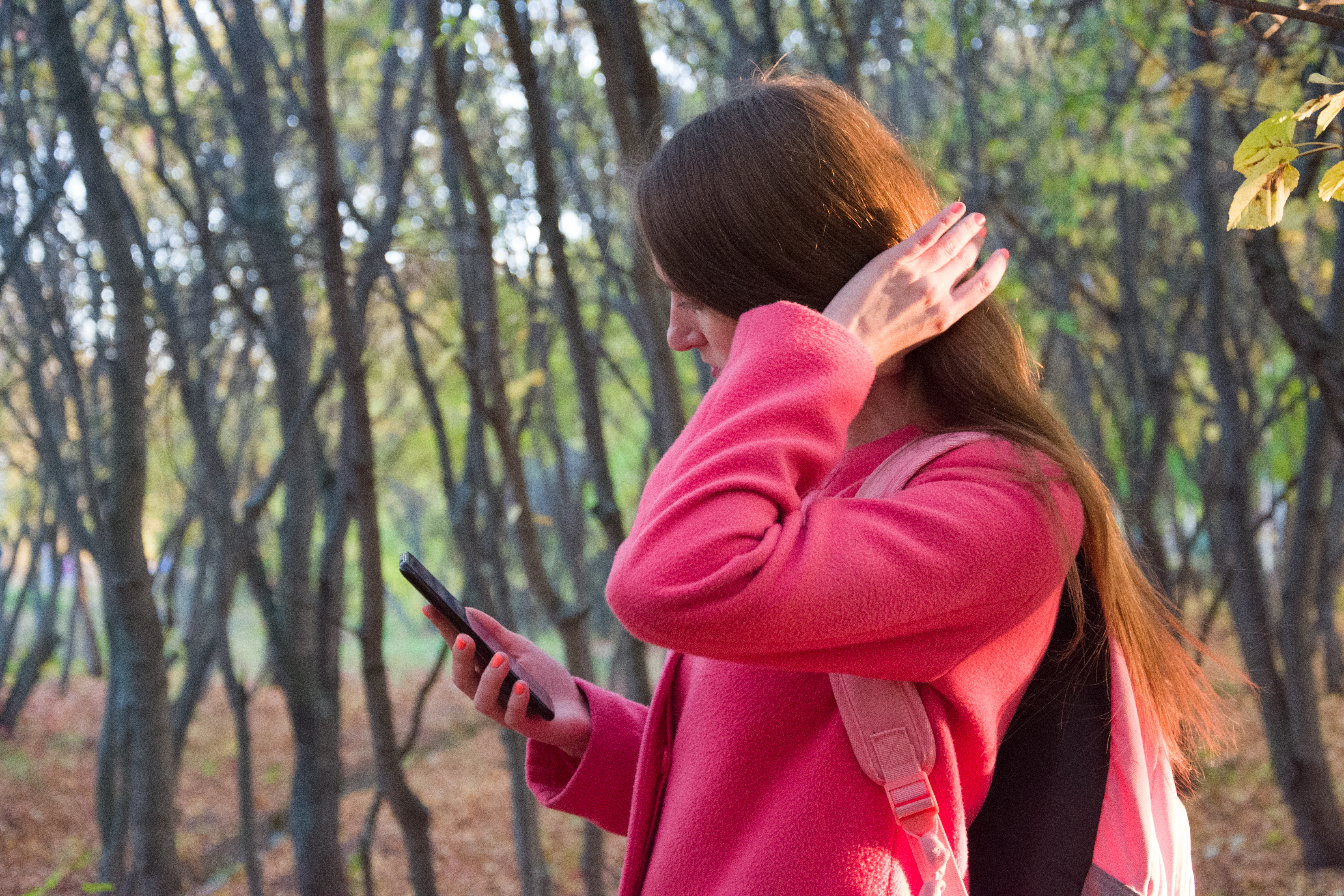 A woman in a pink coat with a backpack uses a smartphone while standing in a wooded area on an autumn day.