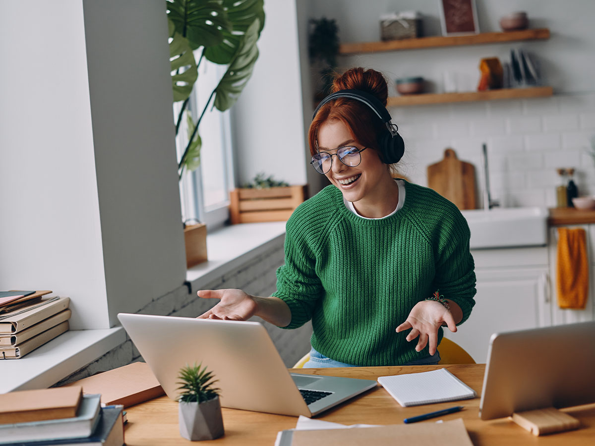 A woman wearing glasses and headphones smiles and gestures while working on a laptop at a home desk, participating in a video call or remote meeting. The workspace features notebooks, a plant, and another laptop, with a bright kitchen in the background.