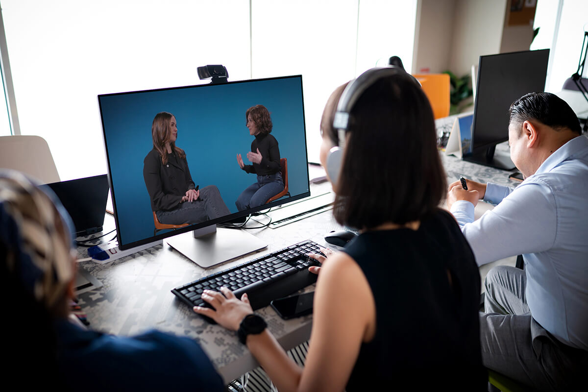 A woman wearing headphones watches a video on a computer monitor in a busy office environment. The video on the screen shows two women having a conversation. Several people are working at desks in the background.