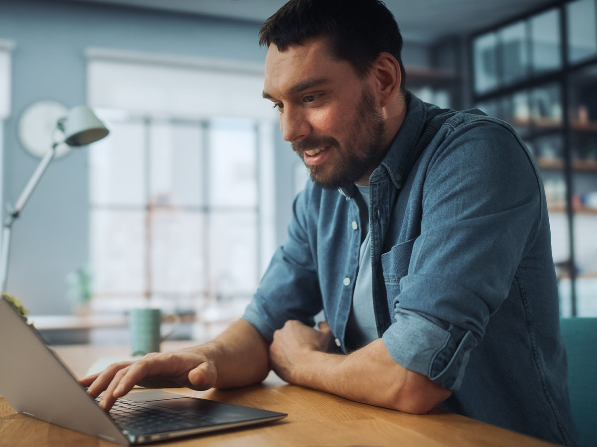 A man wearing a denim shirt is sitting at a desk in a modern home office, working on a laptop and smiling.