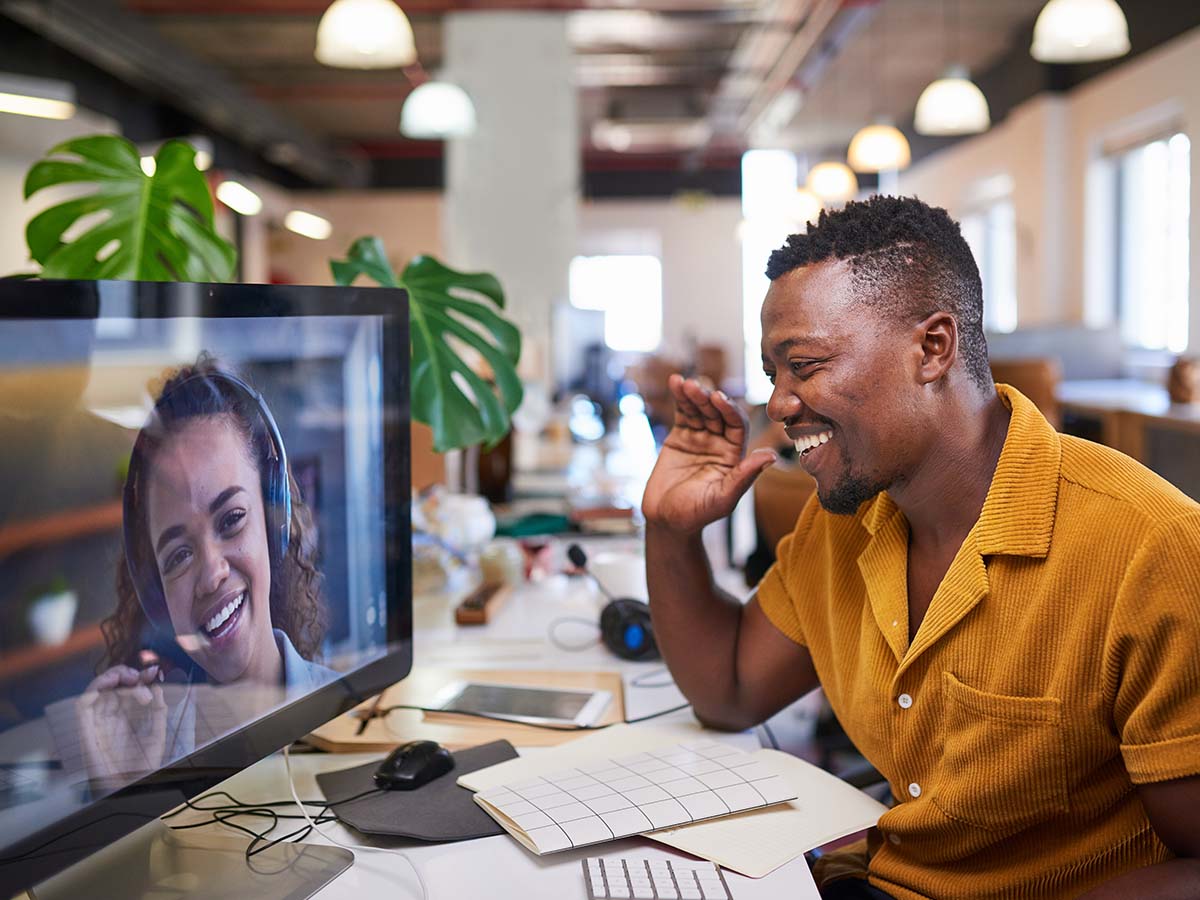 A man sitting at a desk in a modern office waves and smiles at a woman on a computer screen during a video conference call.