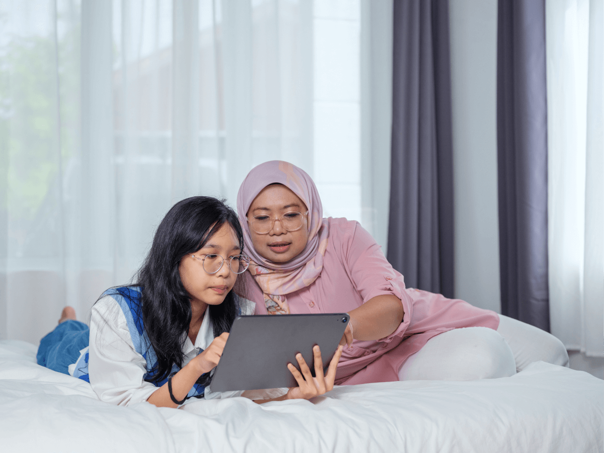 Two people, an adult and a young person, using a tablet together while lying on a bed in a bright room.