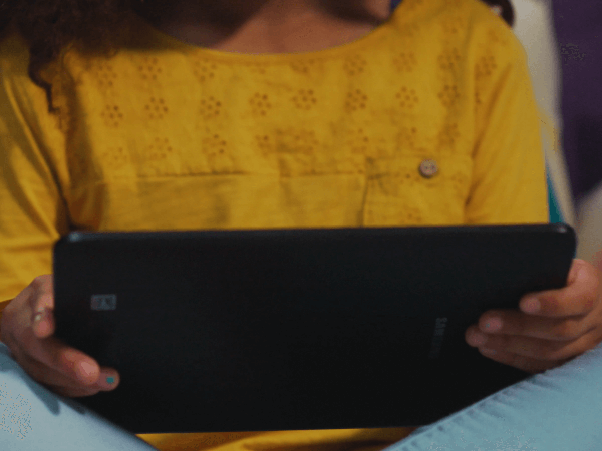 A child wearing a yellow shirt holds a black tablet while seated.