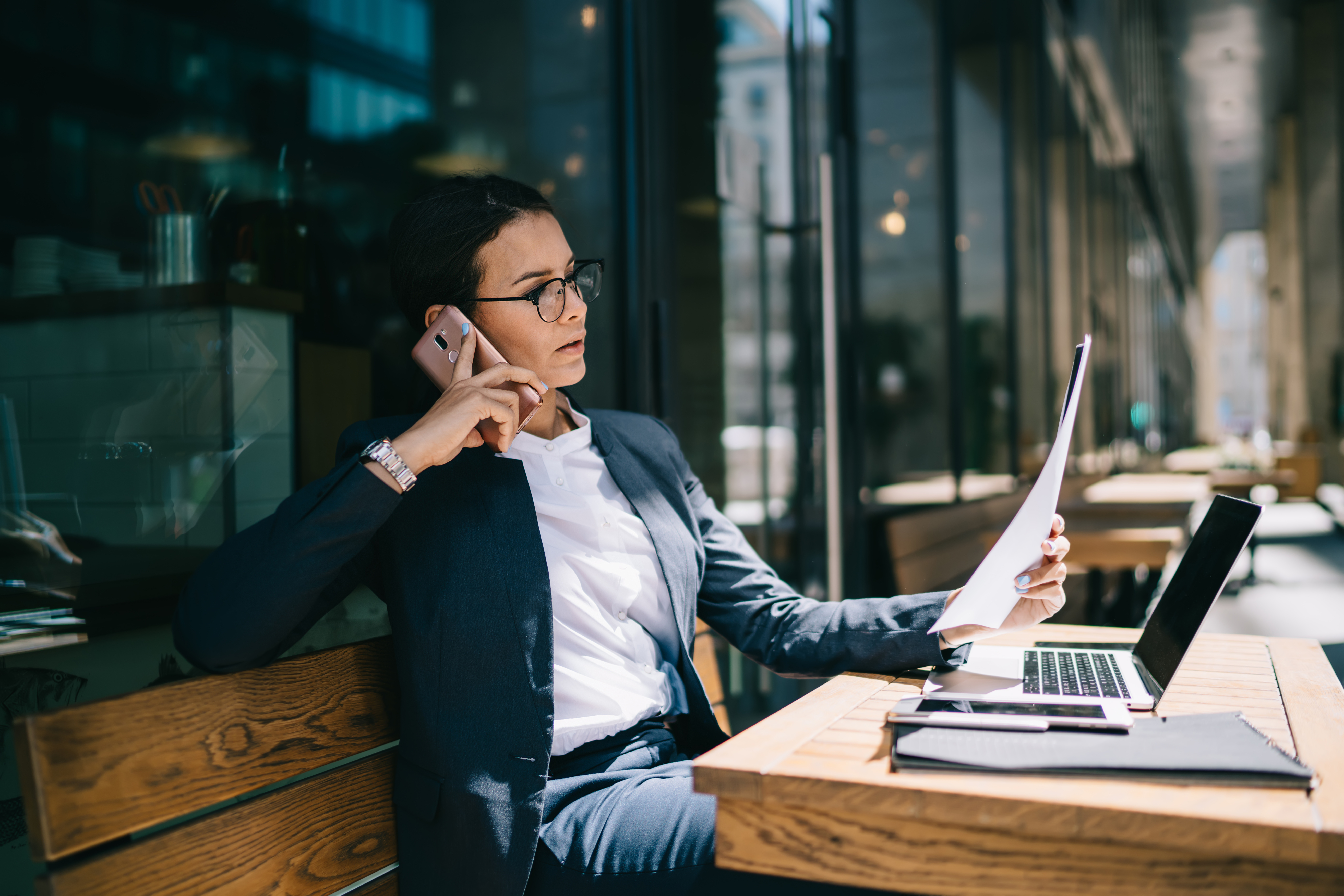 Calm successful trendy woman talking on mobile phone while reading corporate documents during remote job on laptop in cafe patio