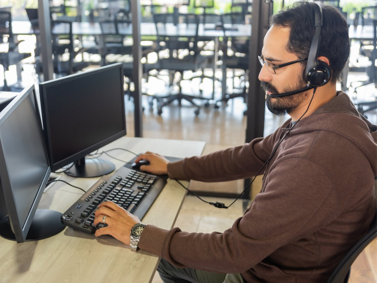 A man wearing a headset is using a desktop computer with dual monitors in a modern office setting.