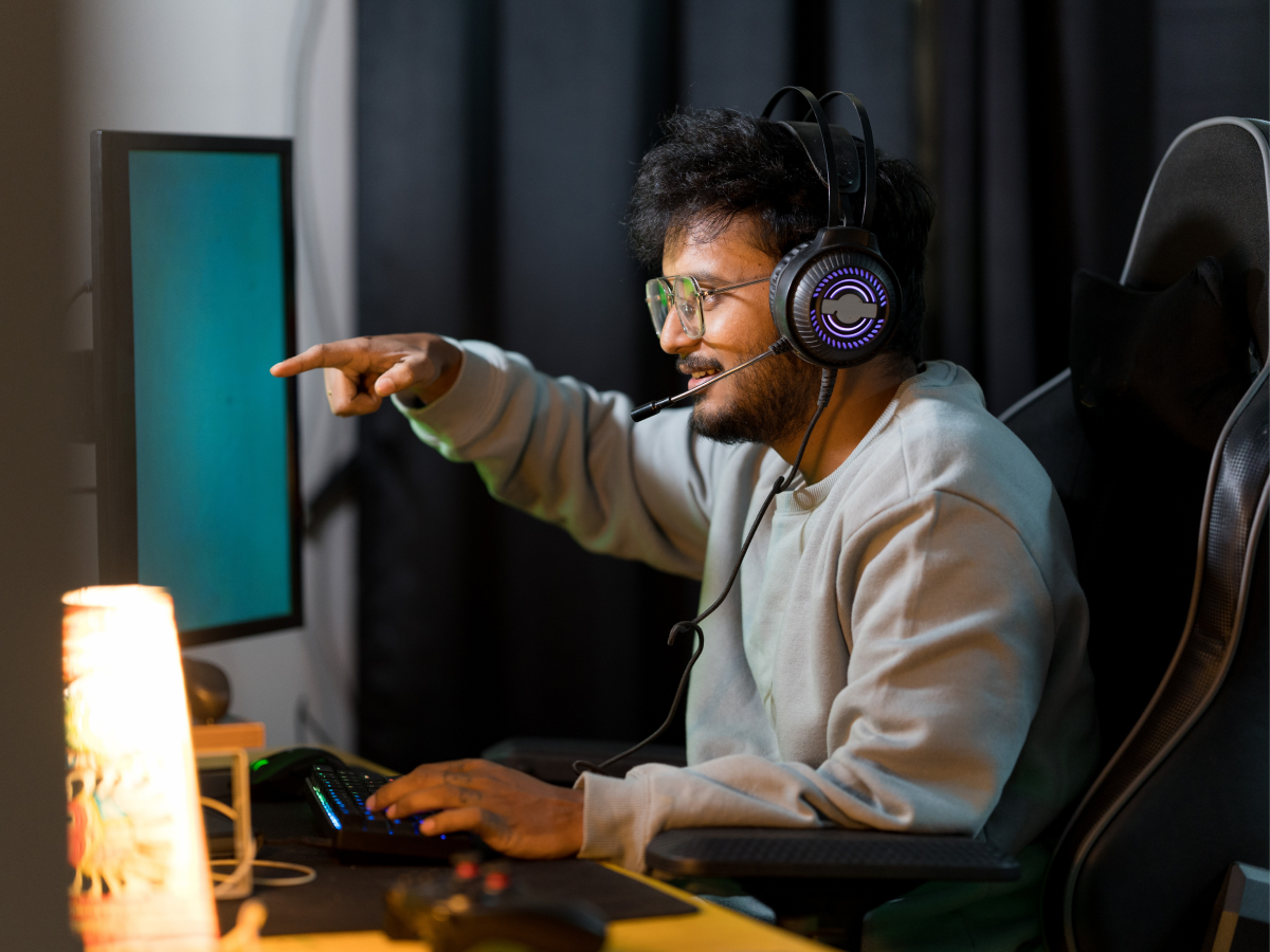 A man wearing a headset and glasses is sitting in a gaming chair, pointing at a computer monitor while using a keyboard, immersed in a gaming session.
