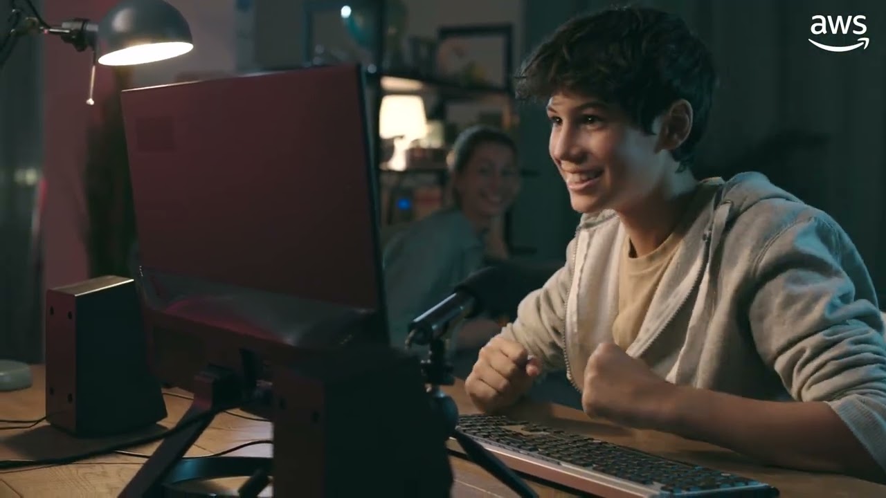 A person sitting at a desk with a computer monitor, keyboard, and microphone, in a dimly lit room with shelves in the background.