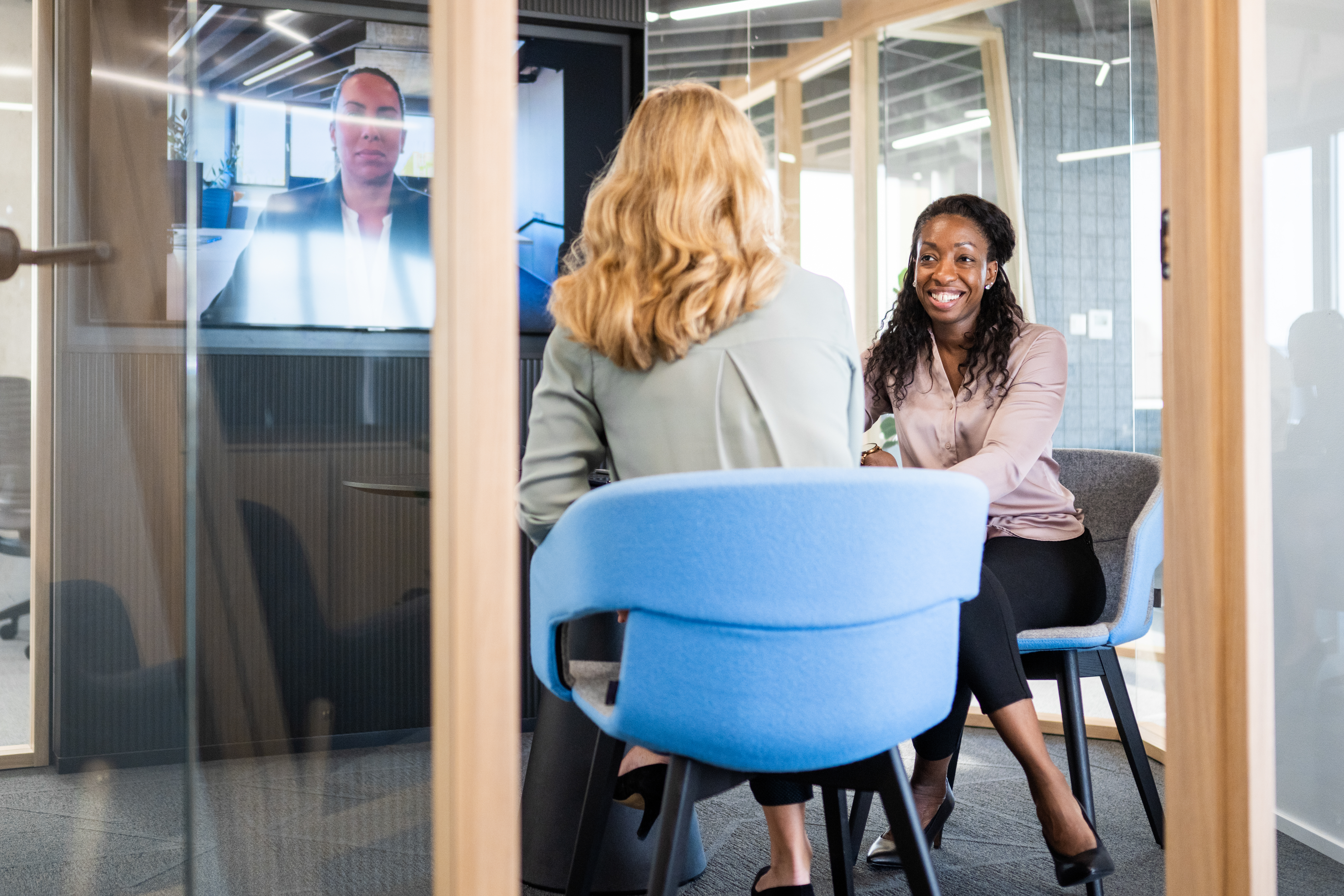 Rear view of two businesswomen attending a video meeting in a small glass conference room.