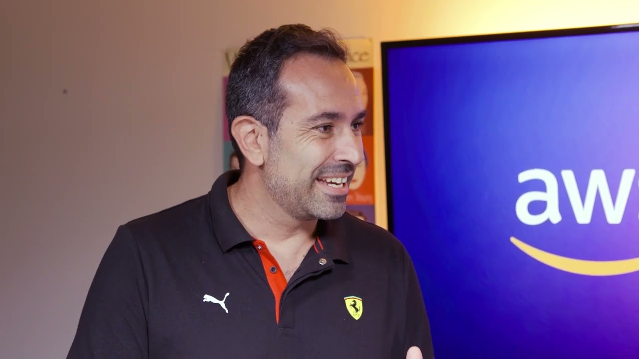 A person wearing a black Ferrari and Puma-branded polo shirt stands in front of a screen displaying the "AWS" logo.