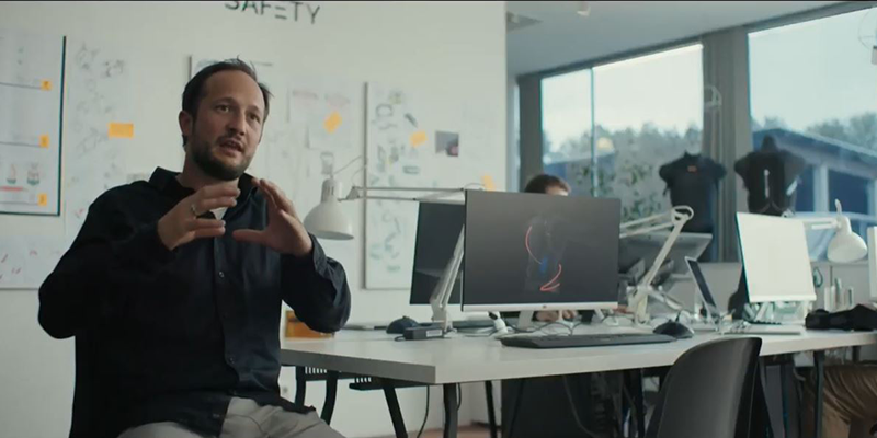 A person is discussing safety or technology inside a modern office space, with desks, computers, and whiteboards in the background. The word 'SAFETY' is visible on the wall, indicating a focus on safety or workplace innovation.