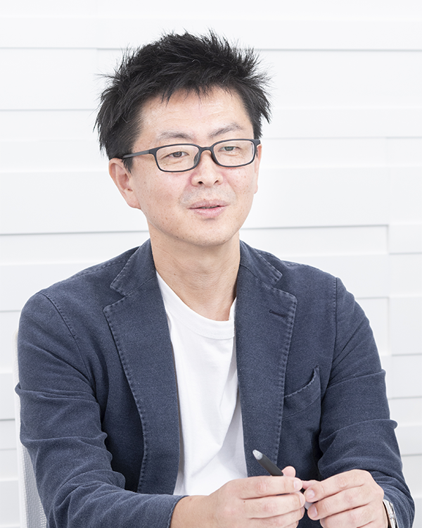 A person wearing a navy blazer and white shirt, holding a pen, seated against a white, textured background.
