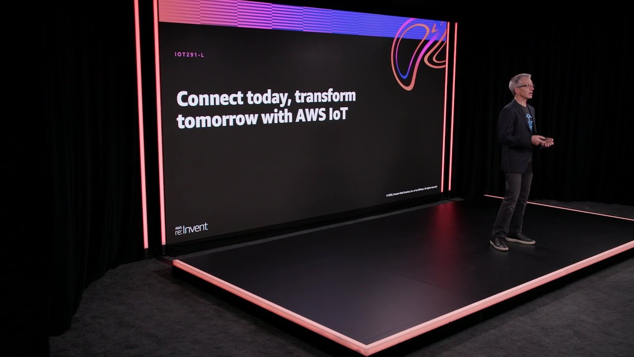 A presenter stands on stage next to a large screen displaying the text 'Connect today, transform tomorrow with AWS IoT' during an AWS re:Invent presentation. The setting is modern with black and pink accents, and the screen features the session code IOT291-L.