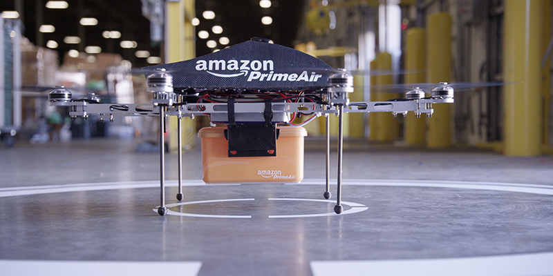 An Amazon Prime Air drone carrying a package, positioned at a landing area inside an industrial facility. The image highlights the use of autonomous drones for delivery in an engineering and industrial environment.