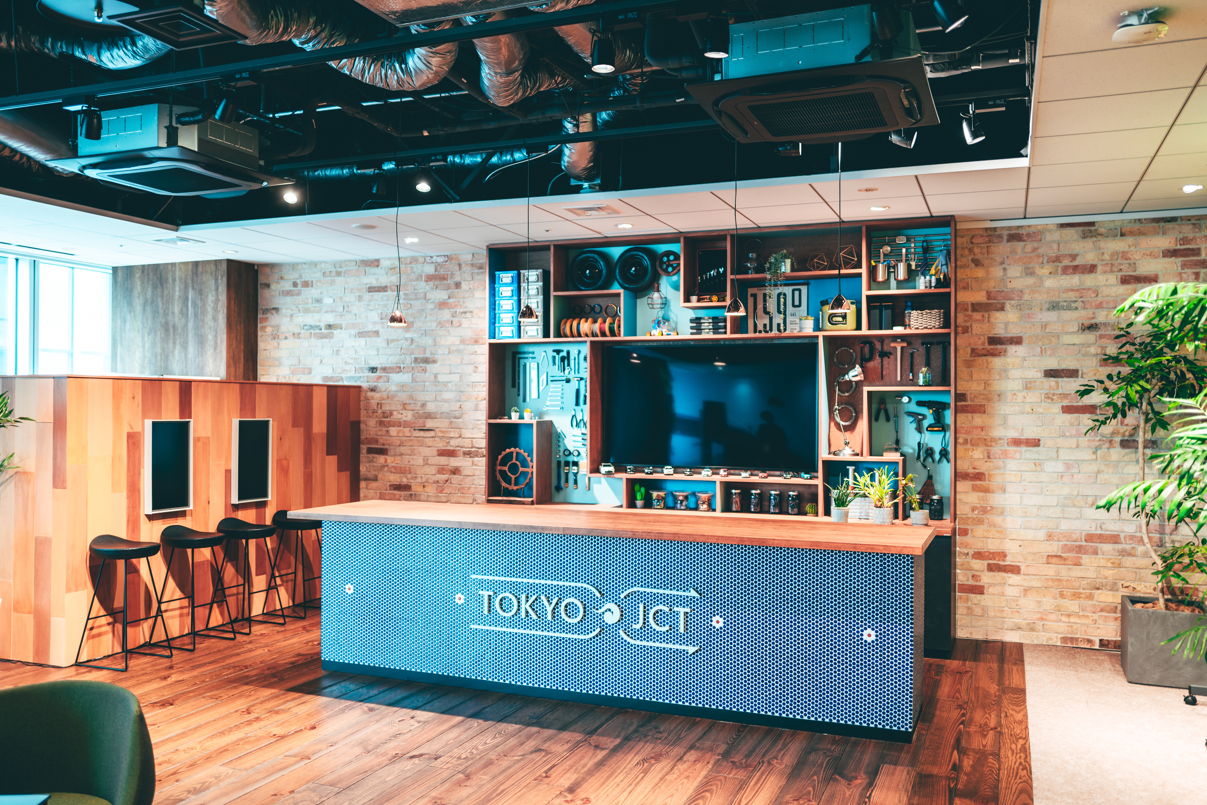 Modern industrial-style workspace with a bar counter labeled "Tokyo JCT," a large TV, shelves displaying tools and decor, and exposed ductwork on the ceiling.