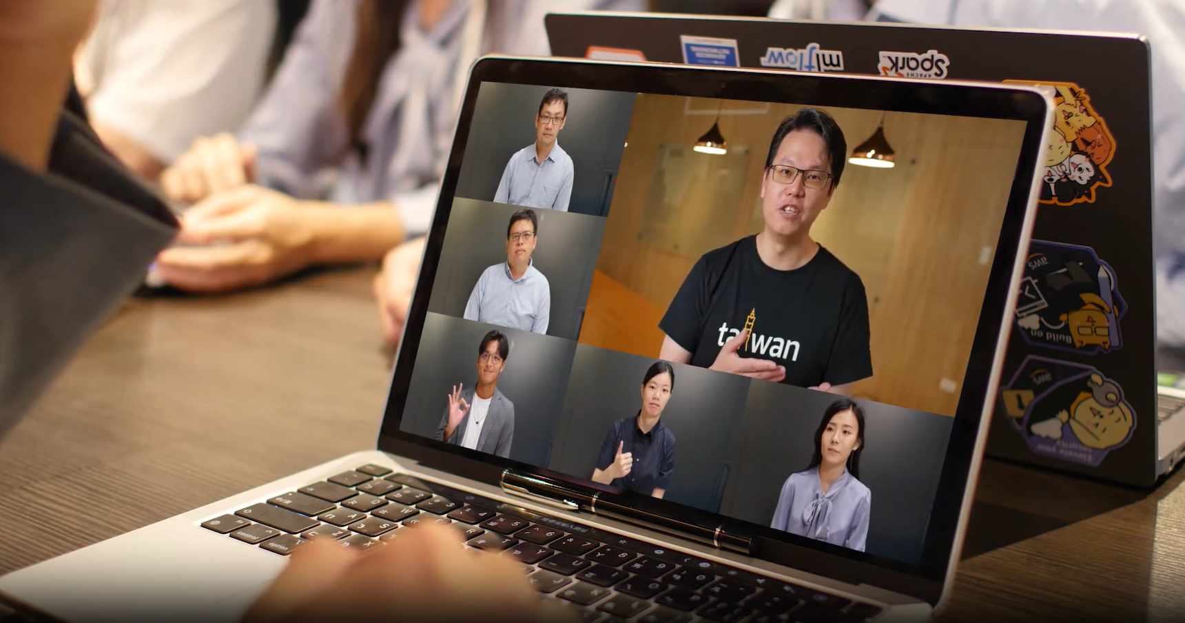 A laptop screen showing a video call with six participants, while hands rest on a table in the background; the other laptop is decorated with colorful stickers.