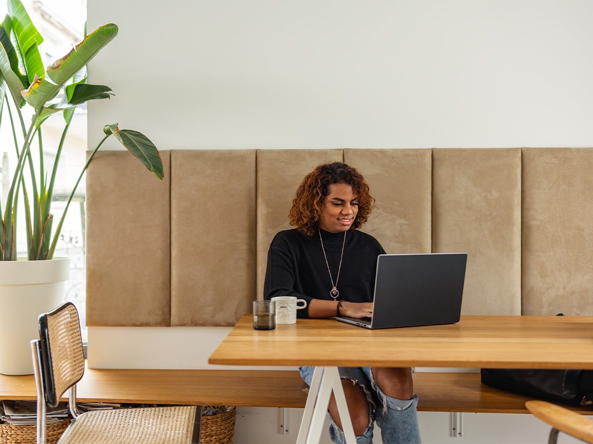 A person sits at a wooden table using a laptop in a modern office or workspace with a large potted plant and beige padded wall panels in the background.