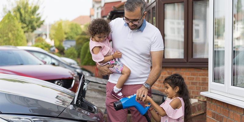 A man holding a child helps another child charge an electric vehicle in a residential driveway, illustrating family engagement with sustainable technology.