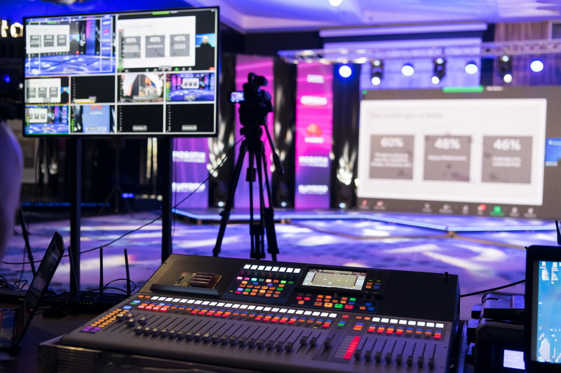 A broadcast control room in a media studio featuring an audio/video mixing console, multiple monitors showing live feeds, a camera setup, and a stage with colorful lighting in the background.