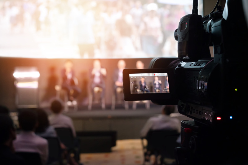 A video camera recording a live panel discussion on stage with an audience in attendance.