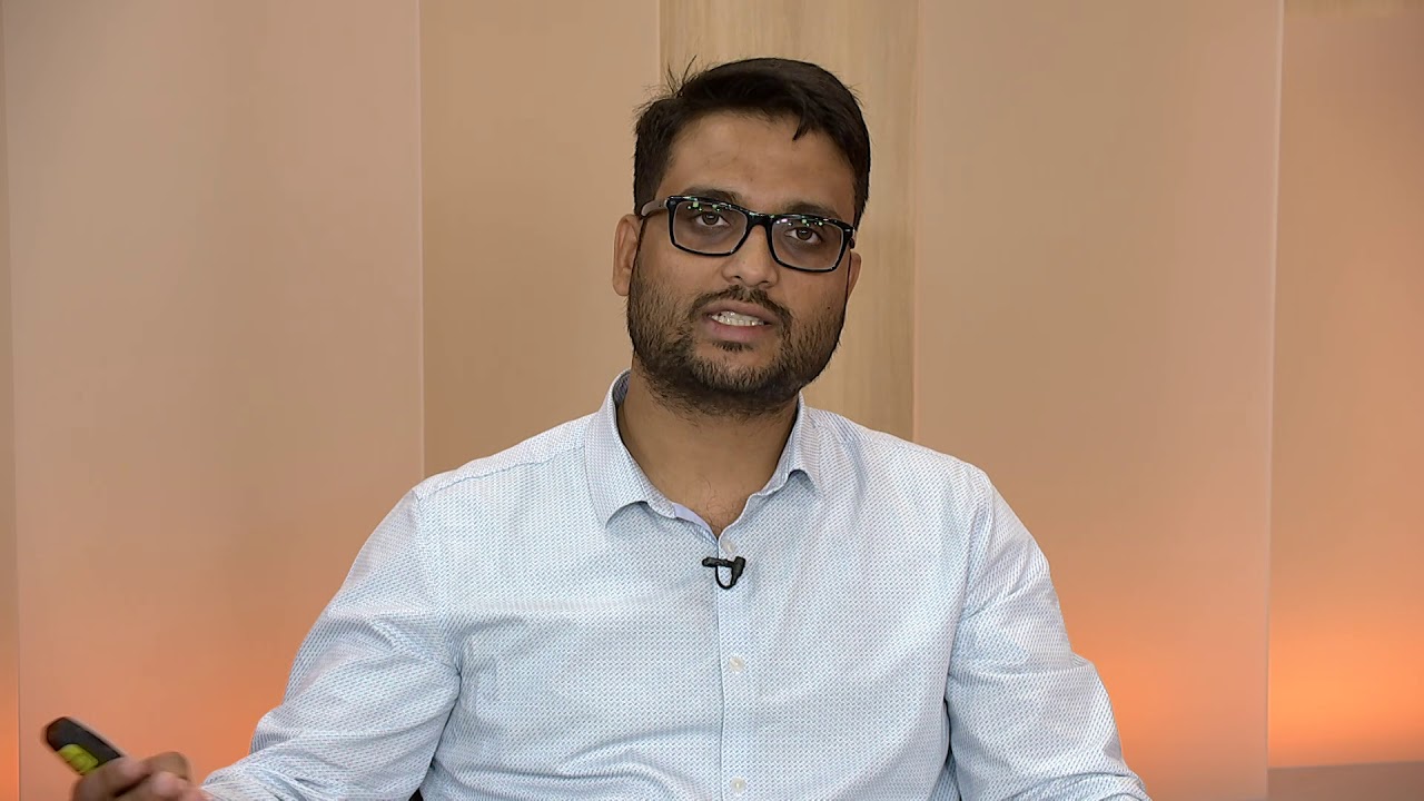 A man wearing glasses and a white shirt is speaking in a studio setting. He has a microphone clipped to his shirt and is gesturing with his hand. The background is neutral with a warm light.