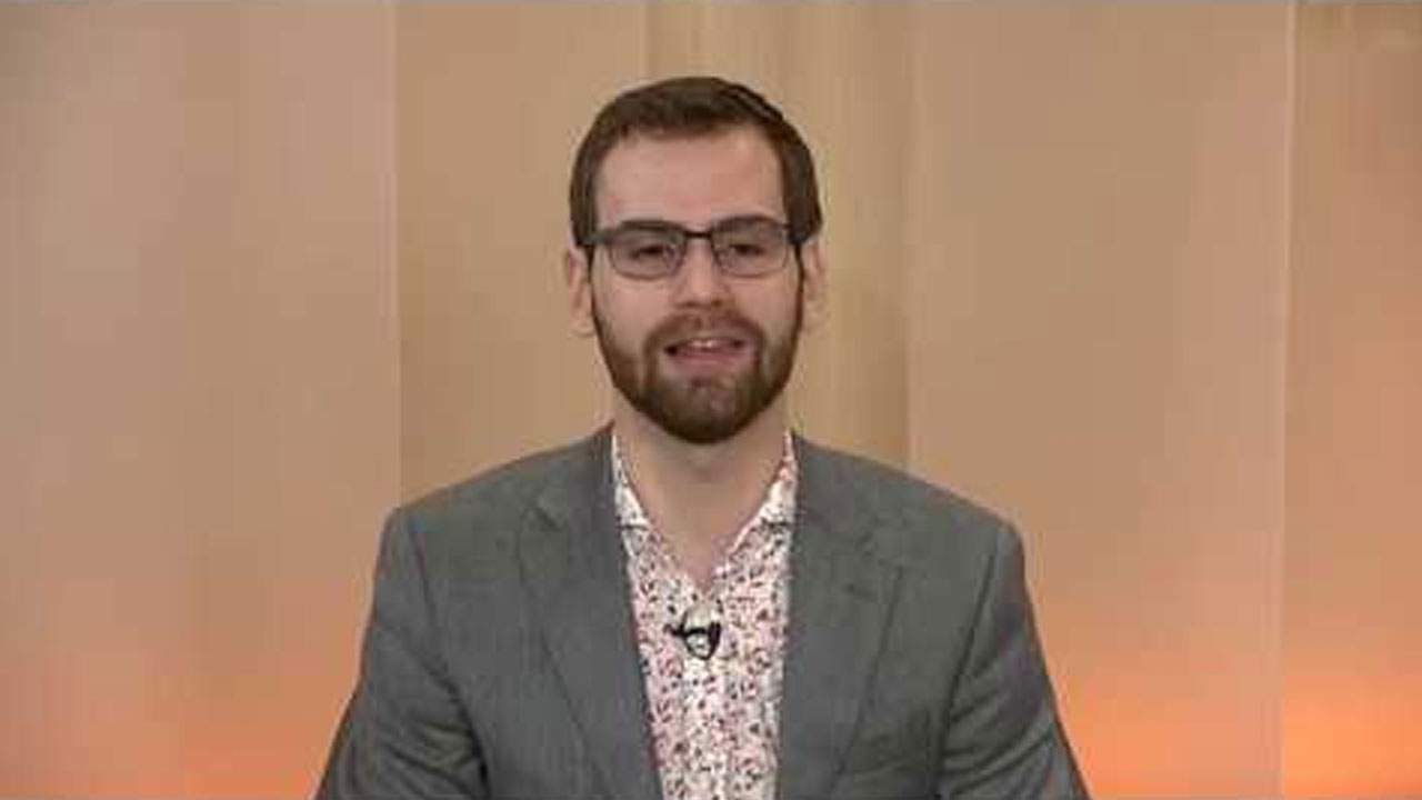 A person wearing glasses and a gray blazer is speaking on camera, against a plain beige background, likely during a presentation or video recording.