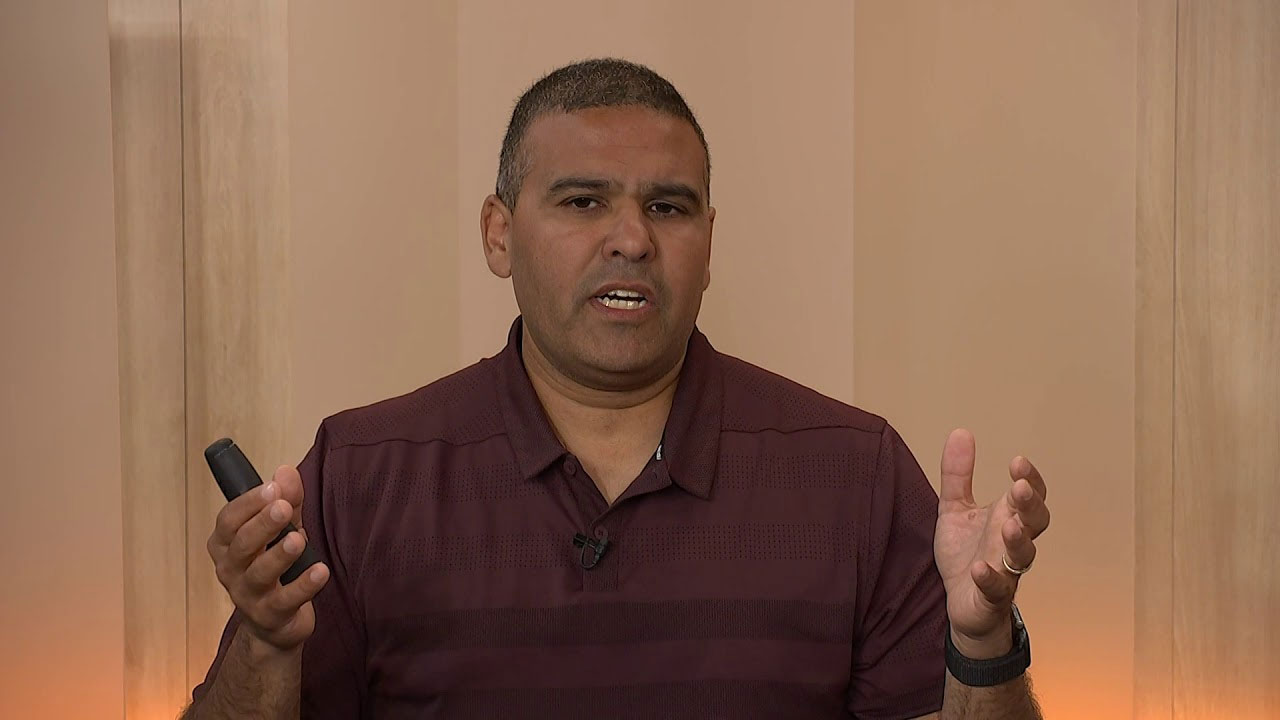 A person in a maroon shirt is giving a presentation or speaking, holding a remote control, in a studio environment.