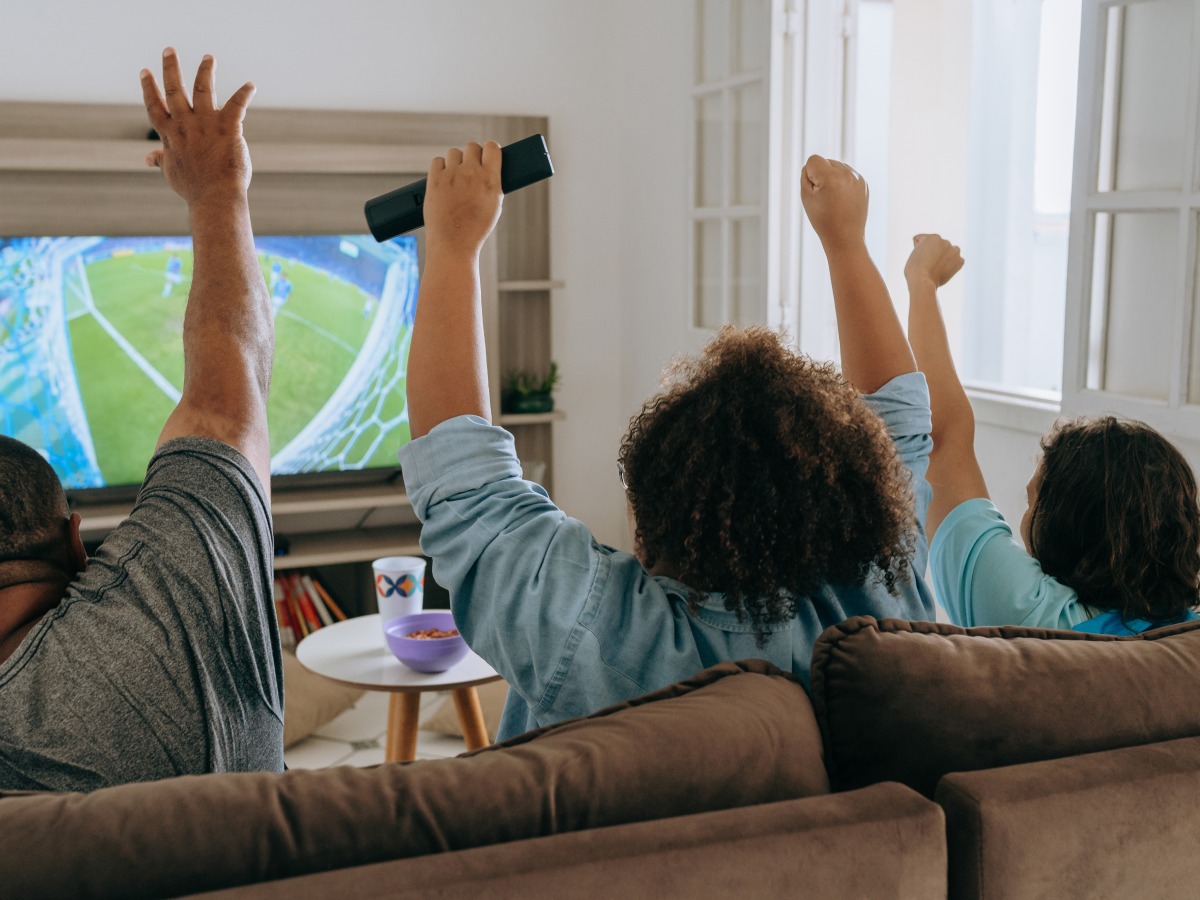 Three people sitting on a couch cheering while watching a soccer game on TV.