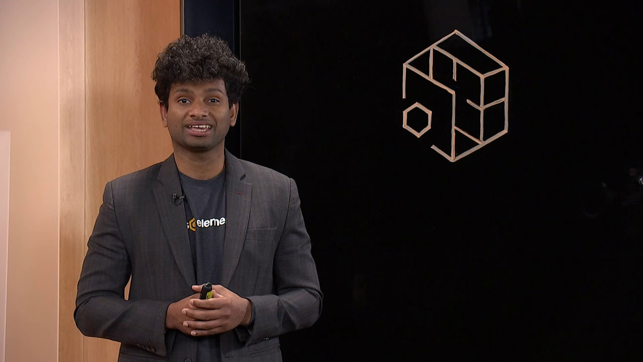 A presenter stands in front of a black screen displaying the AWS Elemental logo. The presenter is wearing a suit jacket and an AWS Elemental t-shirt, and appears to be speaking or presenting in a media services context.