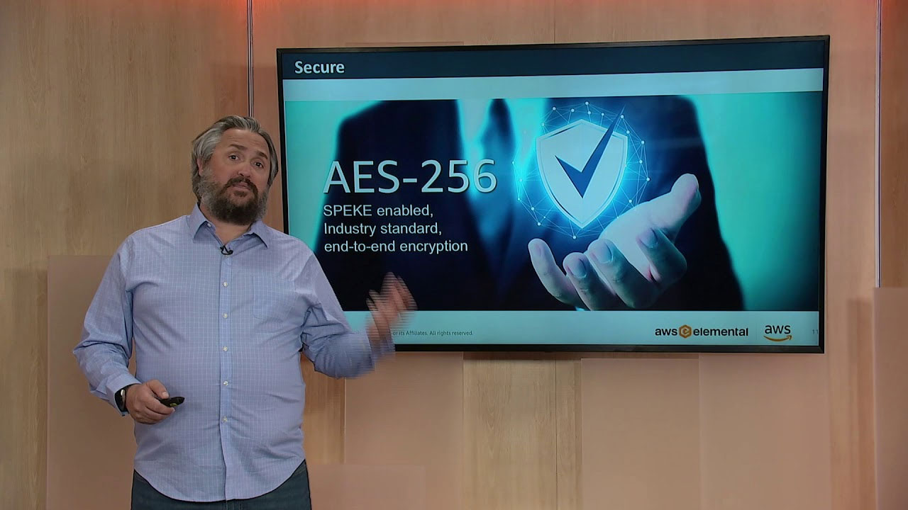 A speaker presenting in front of a screen displaying information about AES-256 encryption, SPEKE-enabled, industry standard, end-to-end encryption for AWS Elemental media services.