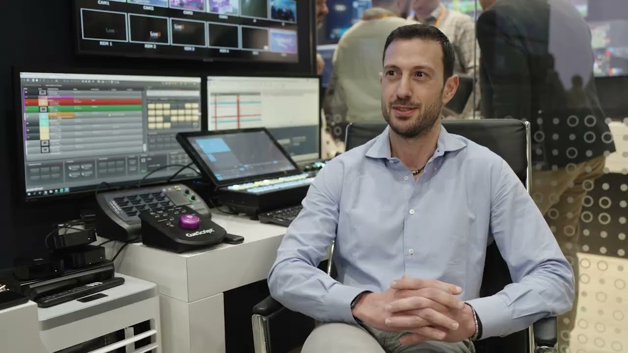 A person sitting in a broadcast video control room with multiple screens and control panels, being interviewed or recorded.