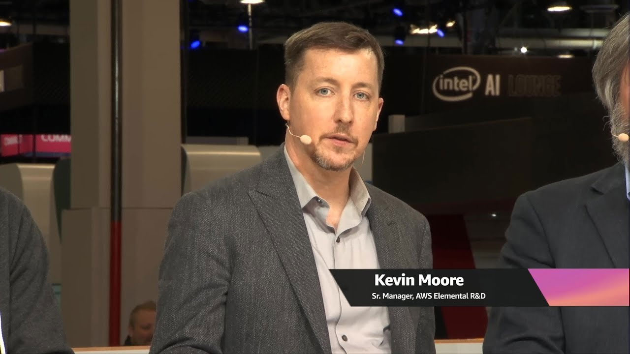 A man speaks during a live AWS Media Services interview or panel discussion at an event, wearing a headset microphone. Event signage and the 'Intel AI Lounge' are visible in the background.