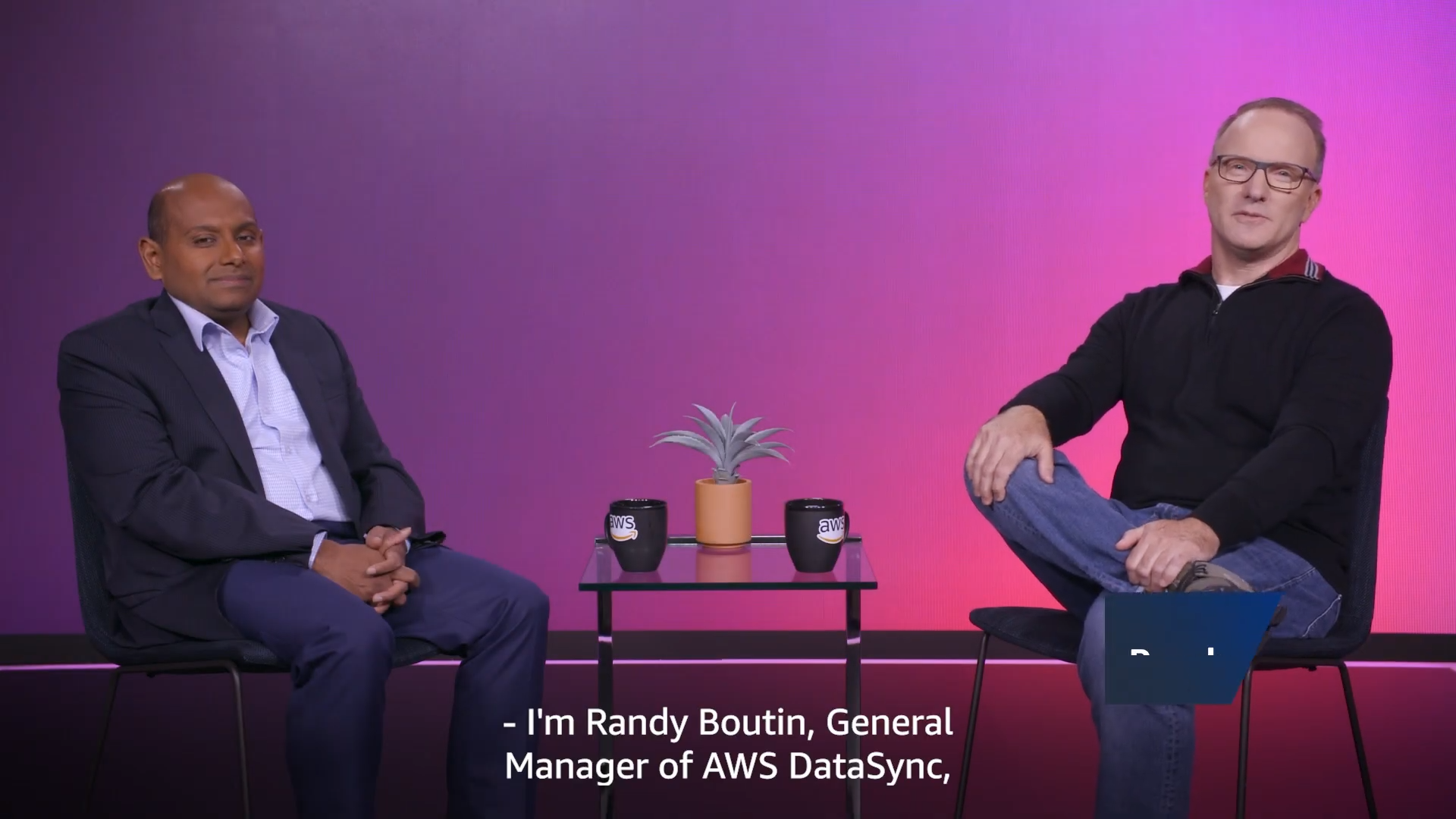 Two men seated for an interview with AWS-branded mugs and a small plant on a table between them, set against a purple gradient background. Subtitle reads, 'I'm Randy Boutin, General Manager of AWS DataSync.'