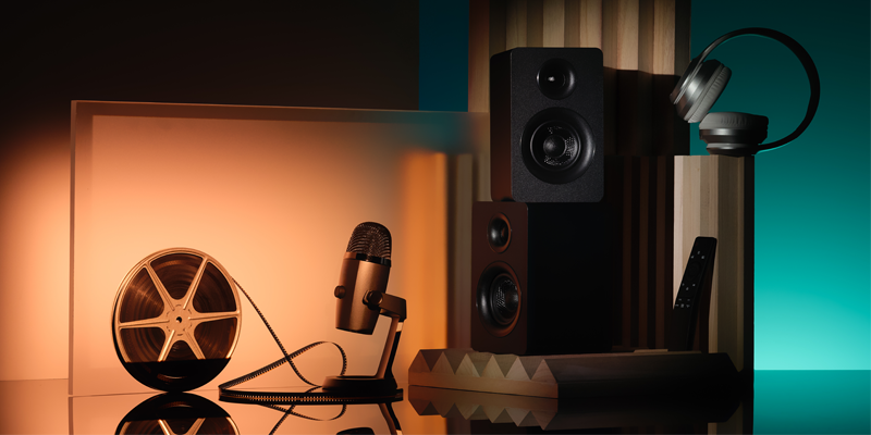 A vintage microphone, film reel, speakers, headphones, and a remote control arranged on a reflective surface with warm and cool lighting.