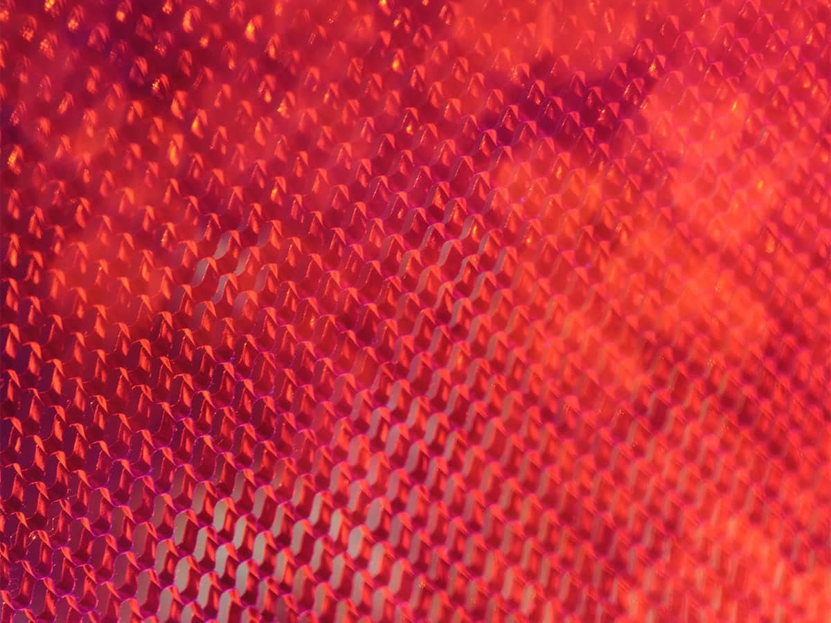 Close-up of a red surface featuring a repeating honeycomb pattern with a geometric, network-like aesthetic. The strong light reflections and vibrant color create an abstract, technology-inspired visual.